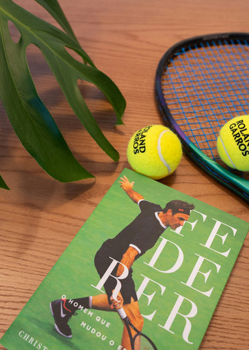 Book about federer, tennis balls, racket, and plant on wood.