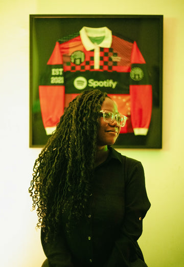 A woman with long dark locs and glasses in front of a framed jersey